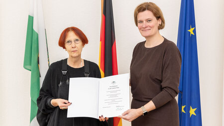 Zwei Frauen, die vor Fahnen stehen, halten gemeinsam eine Urkunde in den Händen. 