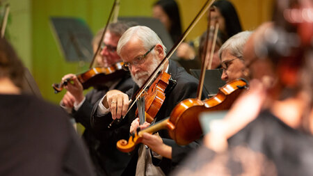 Ein älterer Mann spielt Violine und wird umgeben von weiteren Musikerinnen und Musikern.