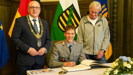 Zwei Männer stehen links und rechts neben einem sitzenden jungen Mann in Uniform, der in ein Buch schreibt.