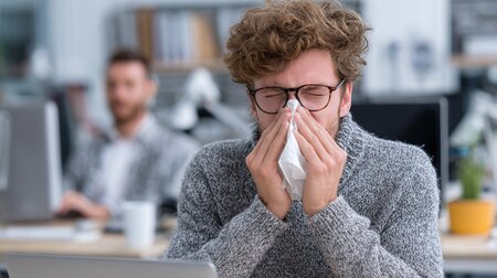 Ein am Schreibtisch sitzemnder Mann schneuzt in ein Taschentuch.