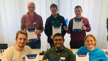 Drei stehende Personen und drei sitzende Personen halten Urkunden in den Händen.