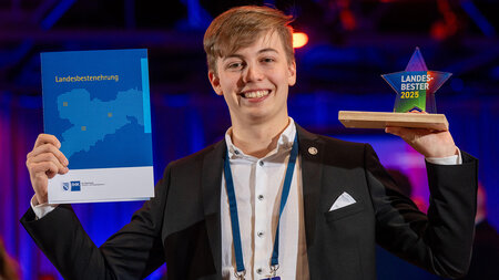 Ein lachender junger Mann hält eine Urkunde und einen Pokal hoch.