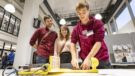 Ein Mann erläutert einem Mann und einer Frau einen Versuchsaufbau.