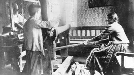 Ein Mädchen und ein Junge arbeiten in einem engen Zimmer gemeinsam mit einem Mann an einem Webstuhl.