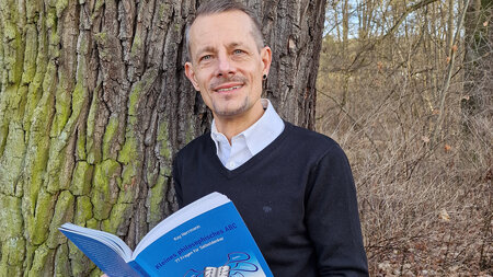 Ein mann steht neben einem Baum und hält ein aufgeschlagenes Buch in den Händen.