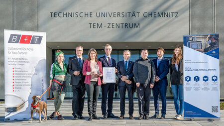 Mehrere Personen stehen nebeneinander vor einem Gebäude. Ein Mann hält eine Urkunde in der Hand.