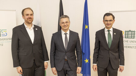 Drei Männer stehen nebeneinander vor Flaggen.