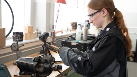 Mädchen steht vor einem Schraubstock und bearbeitet ein Metallstück