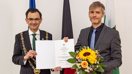 Zwei Männer stehe vor einem Banner und halten eine Urkunde in der Hand