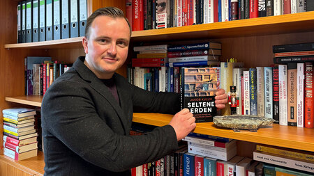 Ein Mann steht an einem Bücherregal und umklammert mit seinen Händen ein Buch.