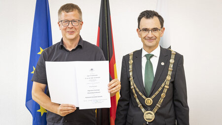 Zwei Männer stehen nebeneinander und halten eine Urkunde in den Händen.