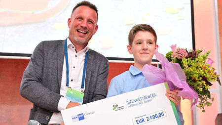Ein Mann überreicht einem Schüler einen Scheck und einen Blumenstrauß.