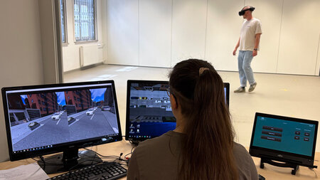 Ein Mann mit VR-Brille auf dem Kopf steht in einem hellen Raum hinter Tischen, auf denen Computer stehen.