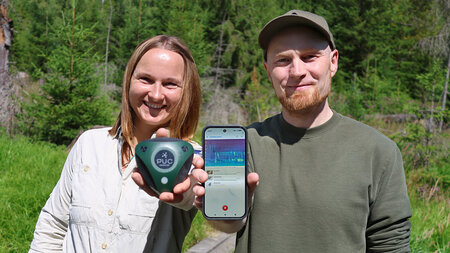 A woman and a man are standing side by side in the woods. She is holding a small electronic device, and he is holding a smartphone.