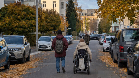 Eine Rollstuhlfahrerin und eine Begleitperson bewegen sich in einem Wohngebiet auf einer Straße..