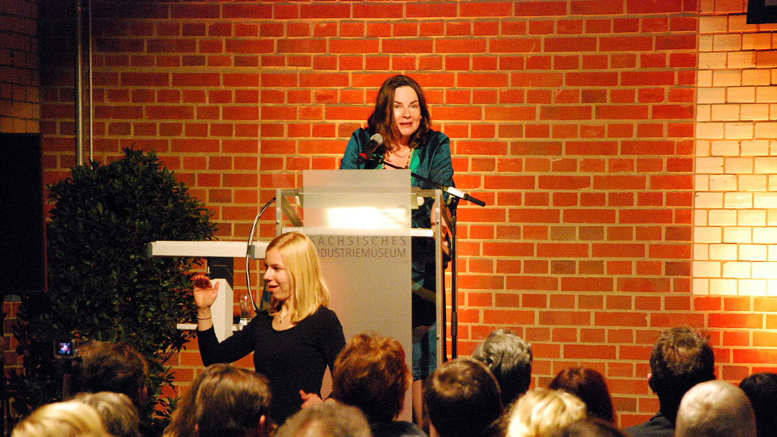 Frau steht vor dem Publikum und gestikuliert, während eine andere am Rednerpult steht und redet
