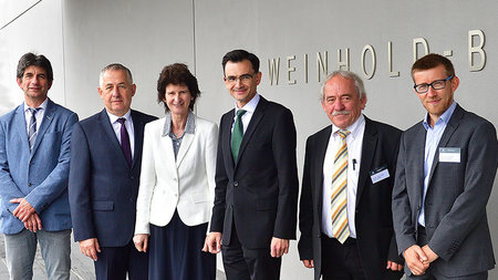 Five men and one woman in suits stand in front of a building labeled “Weinhold-Bau”.