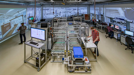 Männer stehen in einer Fabrikhalle und blicklen auf große Monitore an den Wänden der Halle, in der Mitte ist ein Transportband aufgebaut