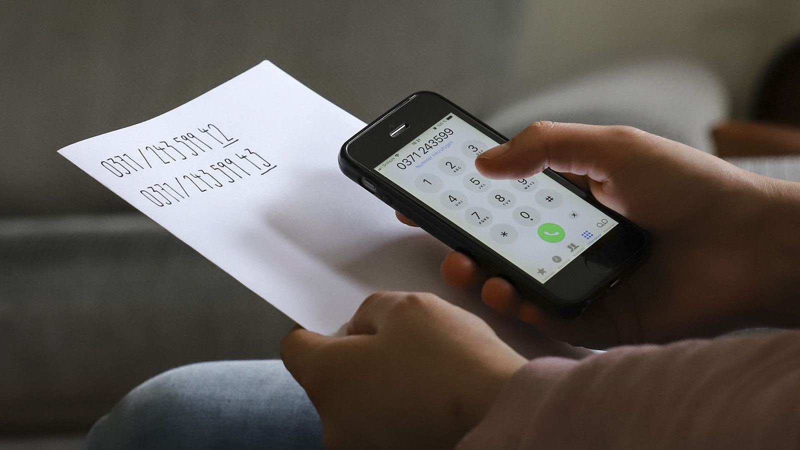 Smartphone in der Hand mit Telefonnumer im Display, daneben zwei Telefonnumern, die auf einem Zettel stehen