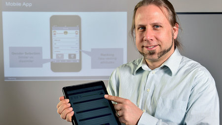 A man in a button-down shirt with a beard holding a tablet computer stands in front of a presentation, which shows a smartphone.