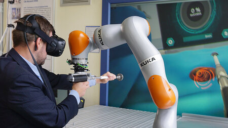 A man wearing a suit and VR goggles holds a drill with help from a robot.