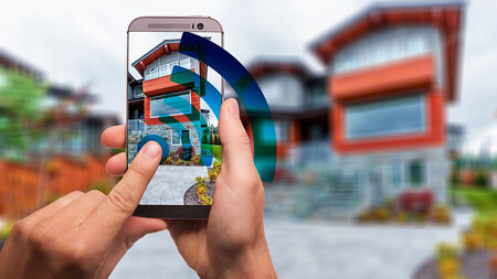 Two hands hold a smart phone, which is taking a photo of a house.
