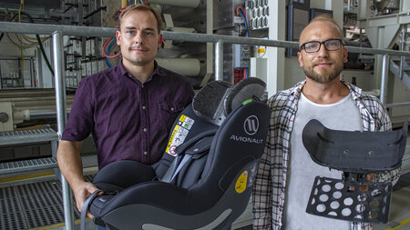 Two men stand in front of a machine and hold a child seat and part of a headrest in their hands.