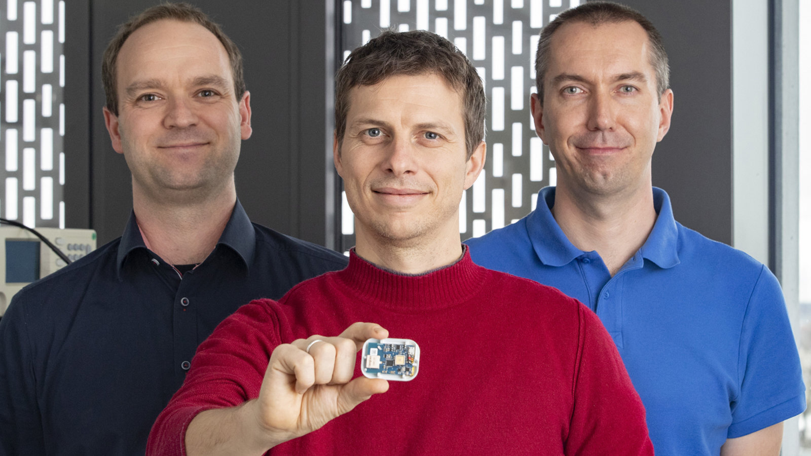 Drei Männer stehen nebeneinander und lächeln. Der Mann in der Mitte hält einen Chip in der Hand.