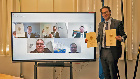 Photo of a man holding up two documents while standing in front of a screen showing a video conference.
