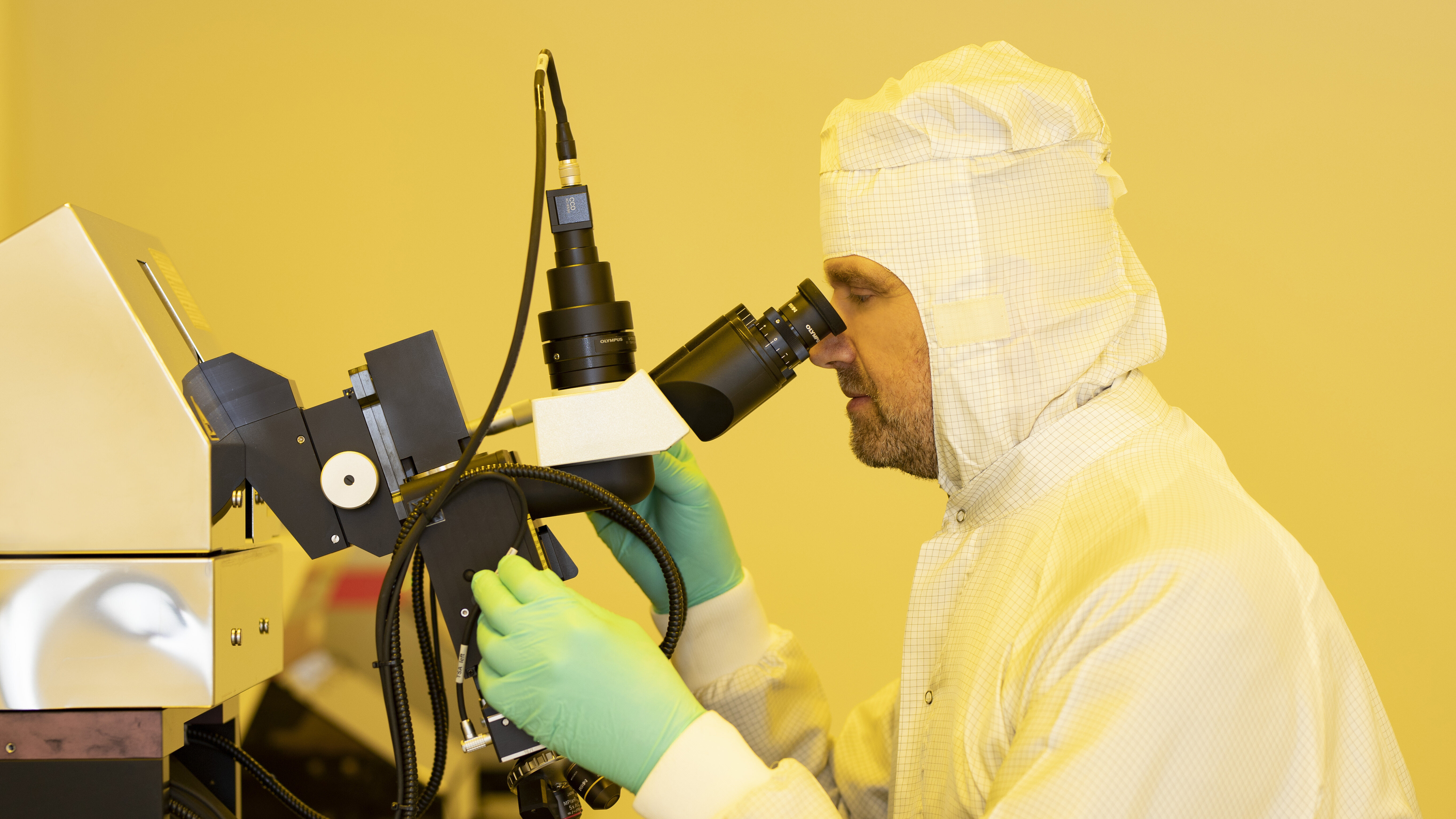 A man in a clean suit sits in front of a microscope