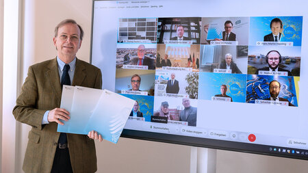 Photo of a man with glasses in a jacket and tie holding three documents while standing in front of a screen showing 14 video conference participants.