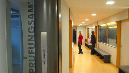 Image of a sign reading “Prüfungsamt” next to two people waiting in a hallway.