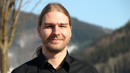 Ein junger Mann mit langen Haaren lächelt leicht.