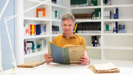Photo of a man holding an old book while sitting at a table in front of a bookshelf.