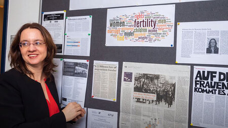 Screenshot of Michèle Tertilt - a woman with glasses and shoulder-length hair standing in front of a bulletin board.