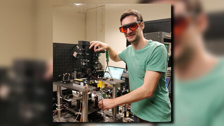 A young man smiles, wears goggles and stand in front of a machine. 
