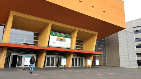 Eine Person läuft in Richtung eines Gebäudes, auf dem Vorplatz sind farbige Abstandsmarkierungen zu sehen.