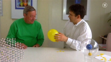 Zwei Männer sitzen an einem Tisch, einer hält einen gelben Luftballon in der Hand,