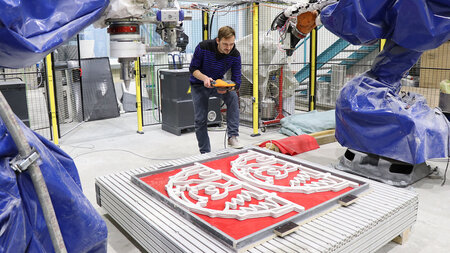 Ein Mann blick auf zwei Schwibbögen, die auf einer roten Matte liegen, links und rechts sind mit Folie ummantelte Roboterarme zu sehen.