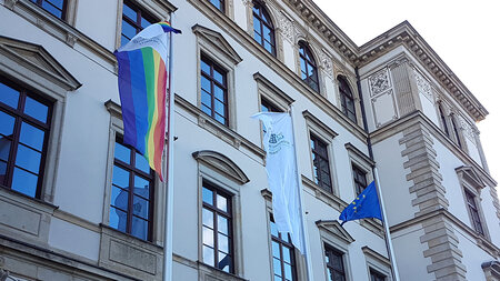 Drei Flaggen wehen an Masten, die vor einem Gebäude stehen.