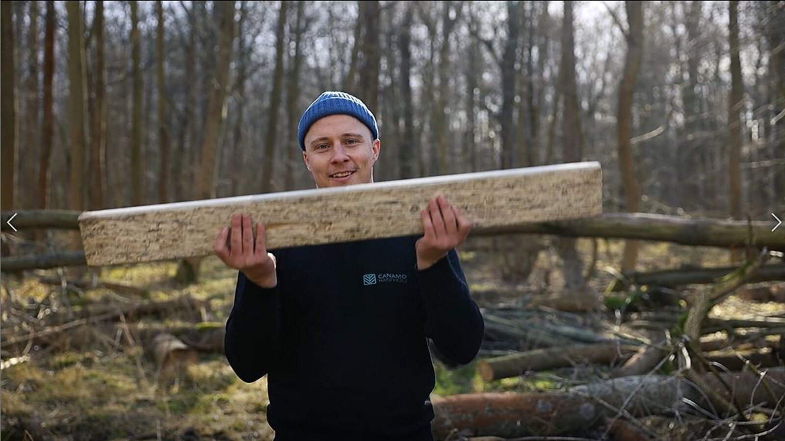 Ein junger Mann mit Mütze steht im Wald und hält ein Stück Holz.