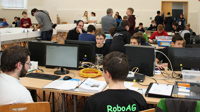 Junge Leute sitzen an mehreren Tischen an Computern und basteln an ihren Robotern.