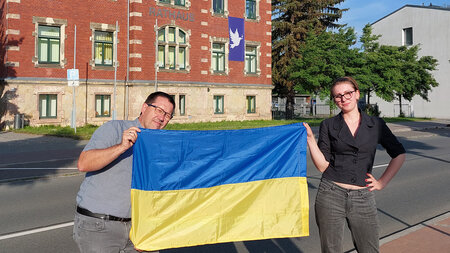 Ein Mann und eine Frau stehen vor einem Gebäude und halten eine blau-gelbe Fahne fest.