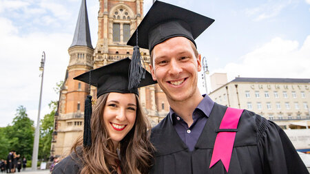 Ein Mann und eine Frau, die in schwarzem Talar und Barett gekleidet sind, stehen vor einer Kirche und lachen. 