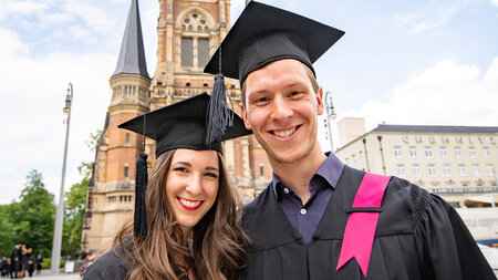 A man and a woman dressed in black gowns and caps standing in front of a church and smiling