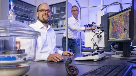 Ein Mann sitzt und ein Mann steht an technischen Geräten im Labor.