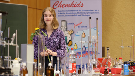 Junges Mädchen mit einer Blume in der Hand steht neben hinter einem Tisch, auf dem viele chemische Versuchsaufbauten stehen. 