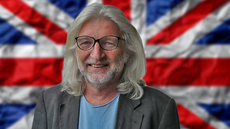 Mann mit Brille und britischer Flagge im Hintergrund.