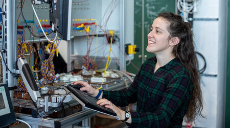 Eine junge Frau mit langen Haaren arbeitet in einem Elektrotechnik-Labor und schreibt auf einer Tastatur.