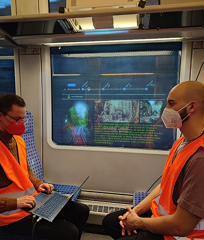 Zwei Männer mit Grsichtsmaske sitzen in einem Zugabteil, einer schreibt an einem Laptop. Am Zugfenster ist eine Projektion zu erkennen.